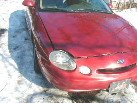 1997 Ford Taurus - A red Ford Taurus is parked on a snowy surface, showing its front end and headlights.