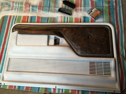 1978 Ford Bronco - Removed white interior door panel with faux-woodgrain trim insert and louvered speaker grille, laid on a striped sheet with hardware.