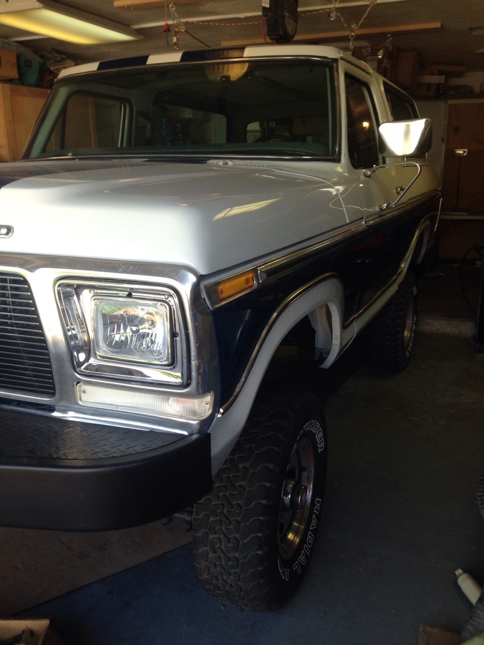 Front 3/4 view of a 1978 Ford Bronco in a garage. It has a two-tone paint job with a dark blue lower half and a silver upper half, with a black and white striped roof