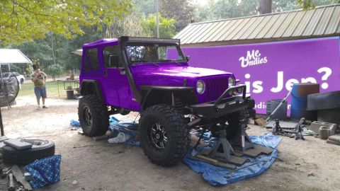 1994 Ford Bronco - A modified purple Jeep Wrangler is elevated on jack stands in an outdoor setting with a large purple banner in the background.