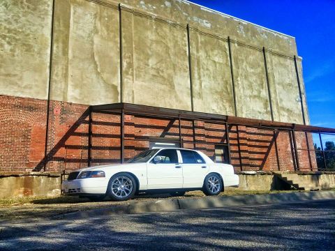 2000 Mercury Grand Marquis - A white 2000 Mercury Grand Marquis is parked next to a weathered brick building under a clear blue sky.