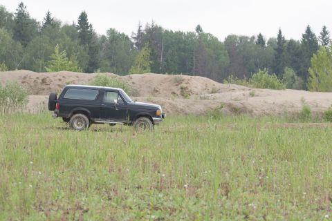 1987 Ford Bronco - 1987 Ford Bronco seen driving through a field with sand dunes and trees in the background.