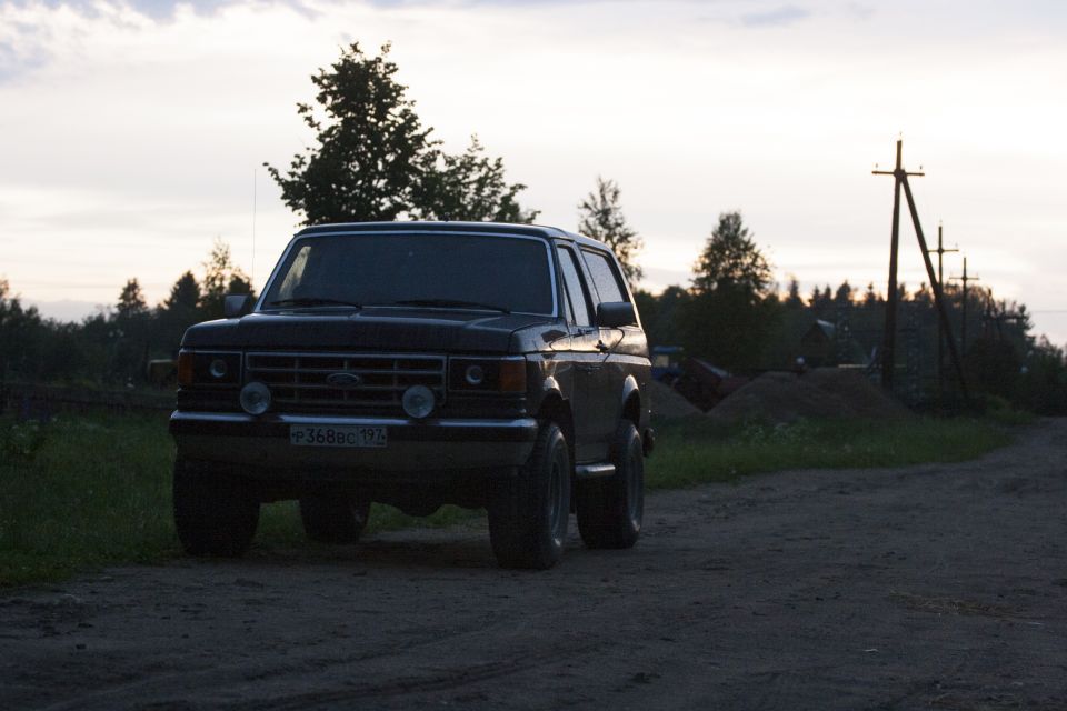 Dark 1987 Ford Bronco parked on a dirt road with trees and utility poles in the background