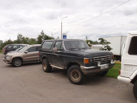 1987 Ford Bronco - A dark 1987 Ford Bronco is parked next to other vehicles under an overcast sky.