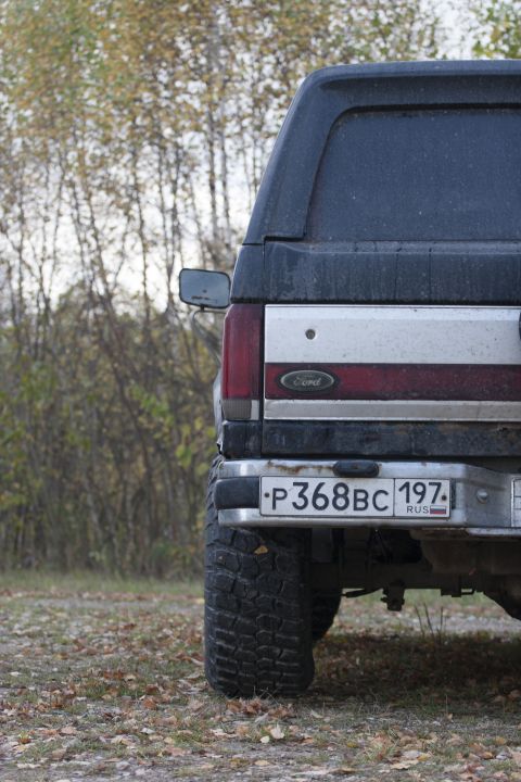 1987 Ford Bronco - The rear of a Ford Bronco is visible, with a dark blue top and silver lower body, set against a backdrop of trees.