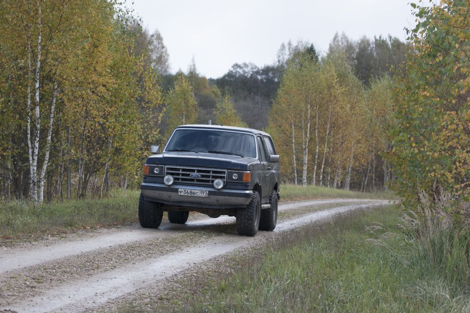 Dark blue 1987 Ford Bronco driving on a dirt road with autumn trees