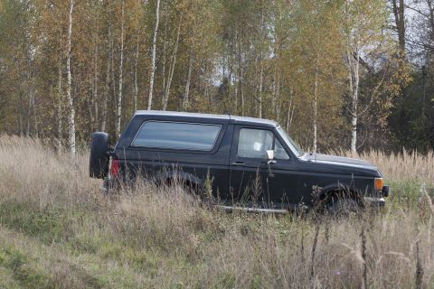1987 Ford Bronco - A black Ford Bronco sits in a field of dry grass, surrounded by trees with autumn foliage.