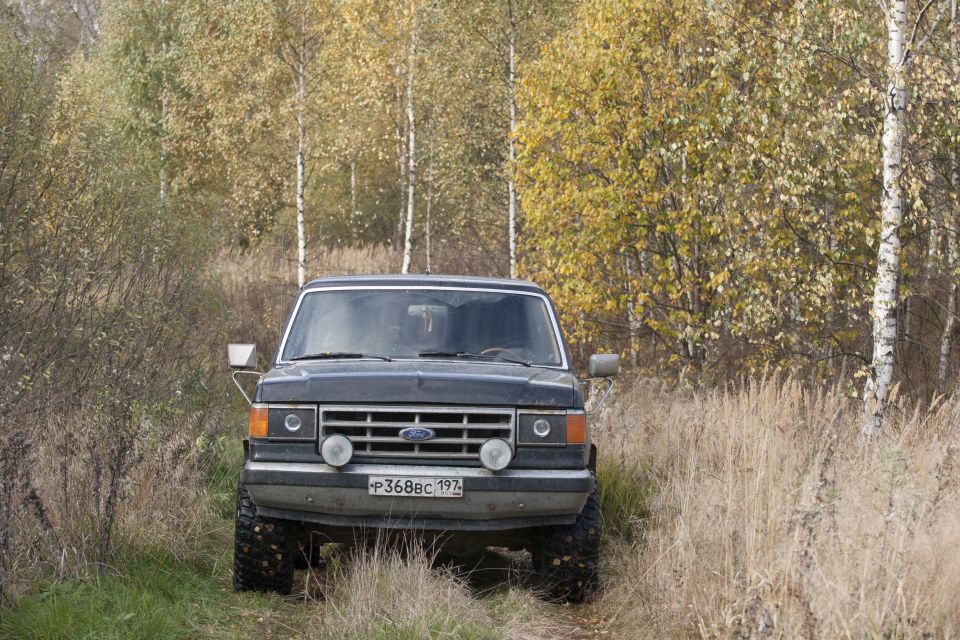 Front view of a dark gray 1987 Ford Bronco parked on a dirt path with dry grass and trees in the background