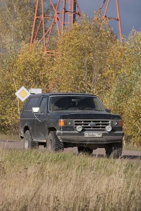 1987 Ford Bronco - 1987 Ford Bronco seen on a dirt road with autumn foliage and a large industrial structure.