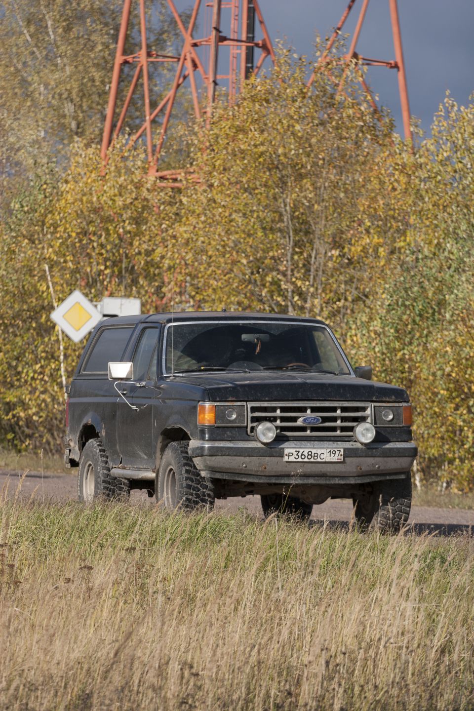 Black 1987 Ford Bronco parked on a dirt road with trees and a large metal structure in the background