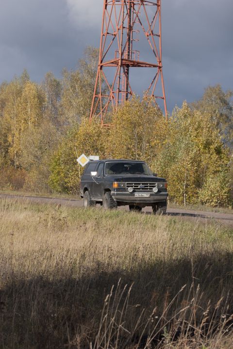 1987 Ford Bronco - A Ford Bronco drives past a tall radio tower with autumn foliage surrounding the area.
