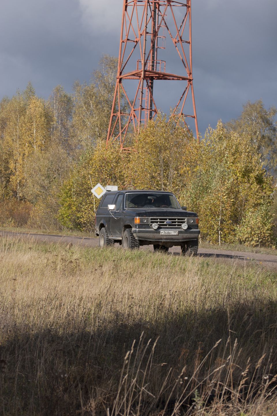 Dark gray Ford Bronco driving on a dirt road near a tall radio tower. Autumn trees in background