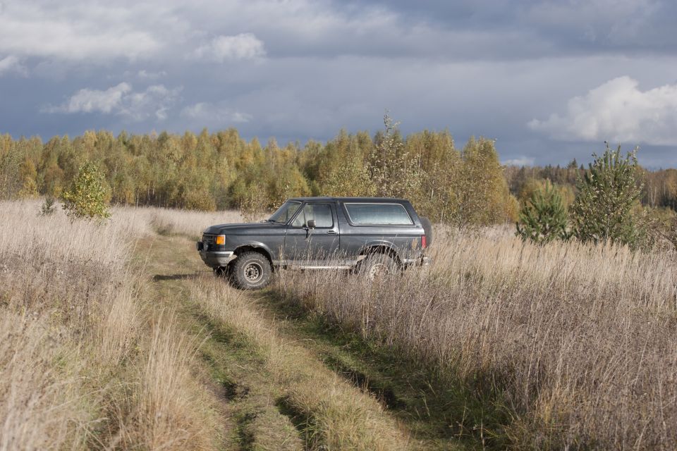 Dark gray 1987 Ford Bronco on a dirt path through tall dry grass, with autumn trees in the background