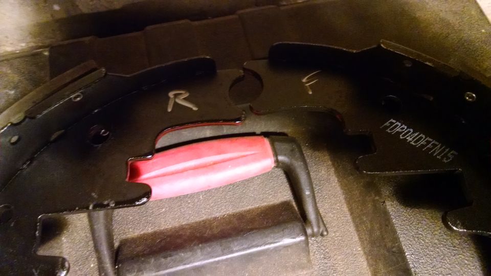 Close-up of two black brake shoes, one marked R and the other F. A red and black tool is between them