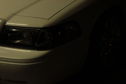 2000 Mercury Grand Marquis - Close-up of a white car's front corner, showing the headlight assembly in shadow.