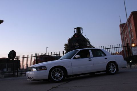 2000 Mercury Grand Marquis - A white sedan is parked near a vintage train and a brick building at dusk.