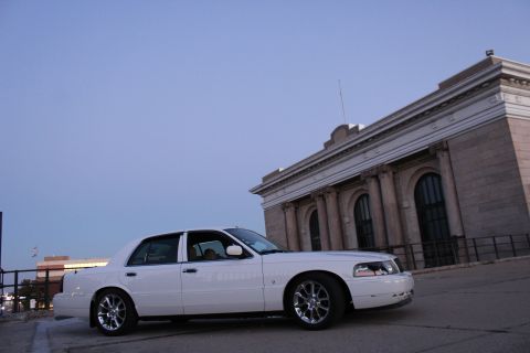 2000 Mercury Grand Marquis - A white 2000 Mercury Grand Marquis sedan is captured from a three-quarter front angle, parked on an elevated surface with a grand building…