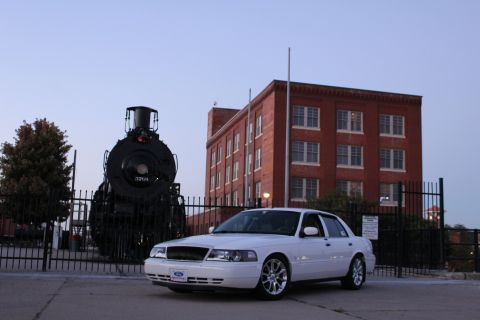 2000 Mercury Grand Marquis - A white sedan is parked in front of a brick building with a steam locomotive on the left.