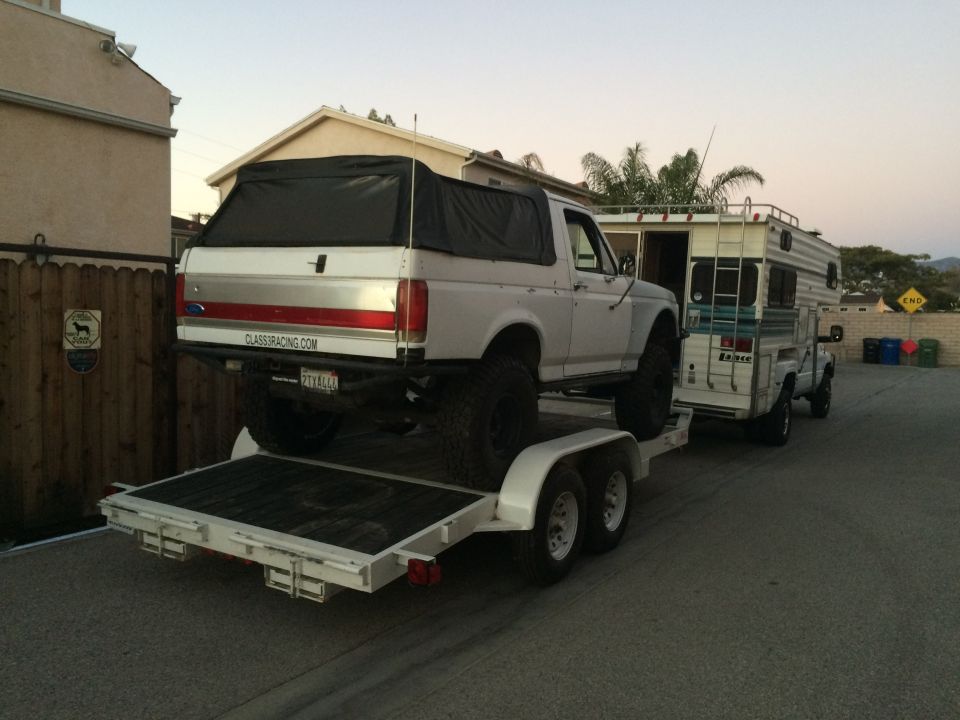 White lifted Ford Bronco strapped to a flatbed car trailer in a driveway at dusk