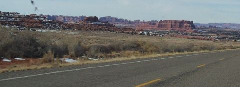 1990 Ford Bronco - A vast desert landscape unfolds under a hazy sky, with distant red rock formations and a paved road in the foreground.