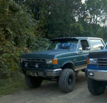 1990 Ford Bronco - A green Ford Bronco is parked on a dirt road next to another vehicle, with trees in the background.