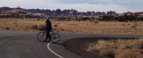 1990 Ford Bronco - Person and bicycle on road in desert with rocky formations. "SUPERMOTORS" logo visible.
