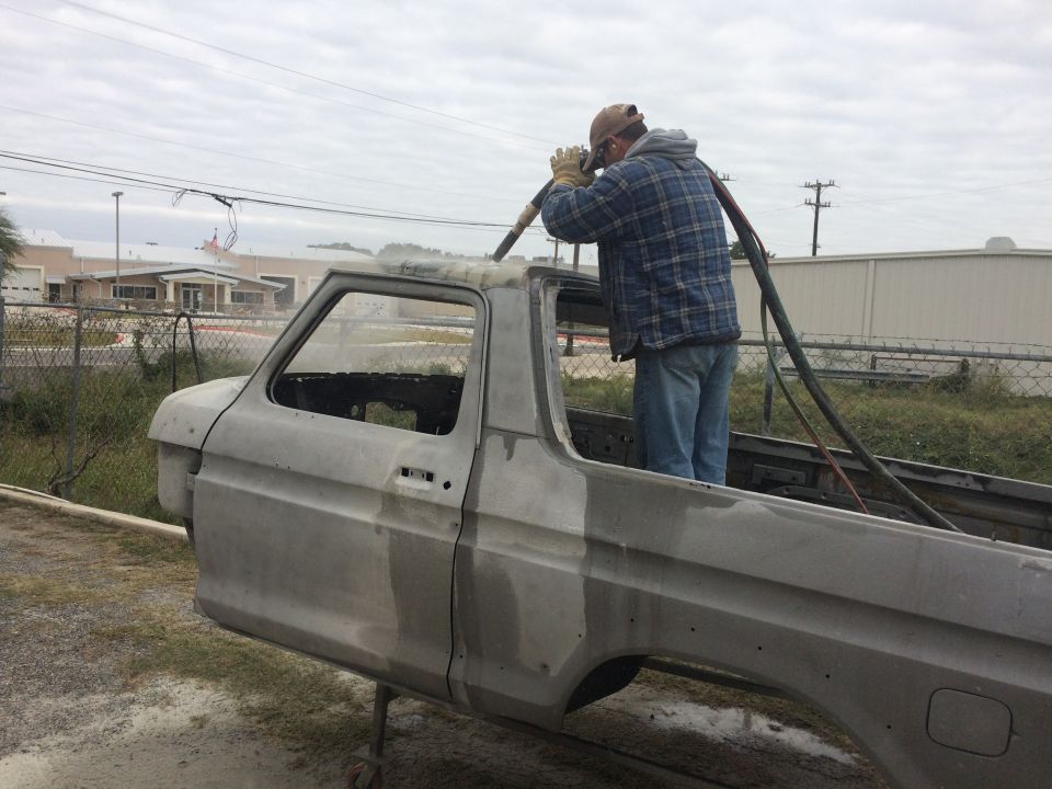 Man media-blasting a stripped gray Ford Bronco body shell on stands outdoors