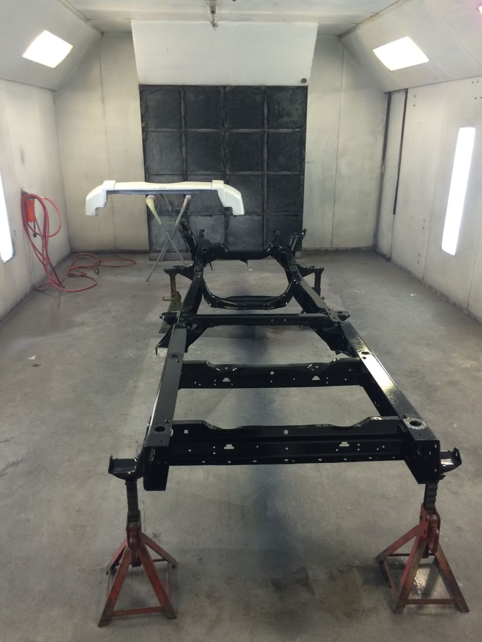 Gloss black Bronco frame on jack stands in a paint booth with a white bumper on a stand behind it