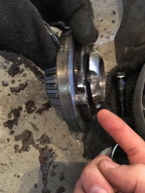 1996 Ford Bronco - A greasy hand points at a tapered roller bearing pressed onto a machined driveline component lying on a concrete shop floor, with a scored,…