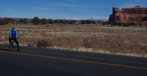 1990 Ford Bronco - A lone cyclist pauses on the side of a road, with a vast desert landscape and distant mountains under a clear blue sky.