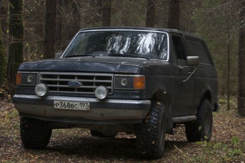 1987 Ford Bronco - A 1987 Ford Bronco sits in a wooded area, its tires slightly muddy from off-road use.