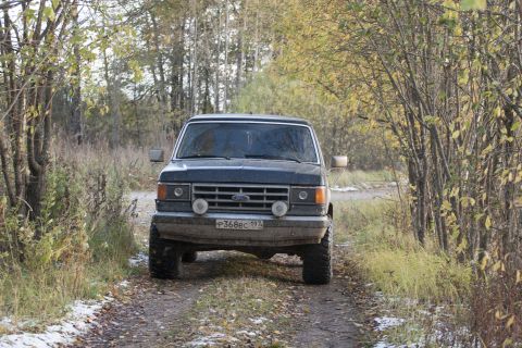 1987 Ford Bronco - A dark-colored Ford Bronco is parked on a dirt road surrounded by trees in autumn.