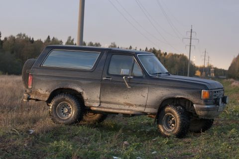 1987 Ford Bronco - A muddy 1987 Ford Bronco is parked off-road in a grassy field with trees in the background.