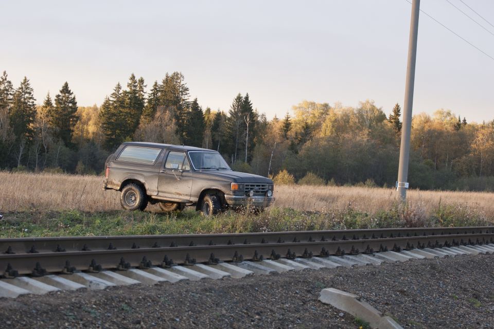 1987 Ford Bronco parked in a field beside railroad tracks, with trees in the background