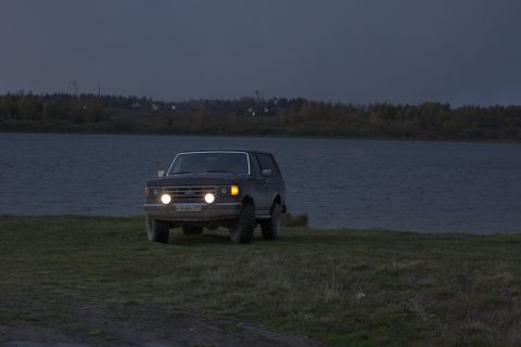 1987 Ford Bronco - An SUV is seen from the front with its headlights illuminated, parked on a grassy area next to a body of water as twilight descends.