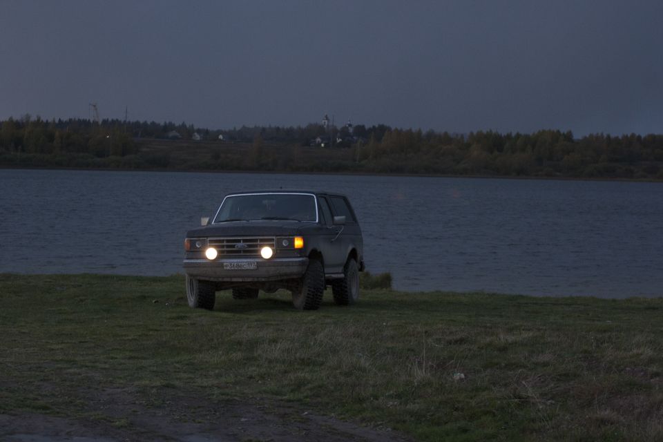 SUV with headlights on, parked near water at dusk