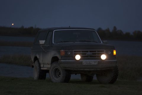 1987 Ford Bronco - A dark-colored SUV is seen from the front with its headlights illuminated against a dusky sky.