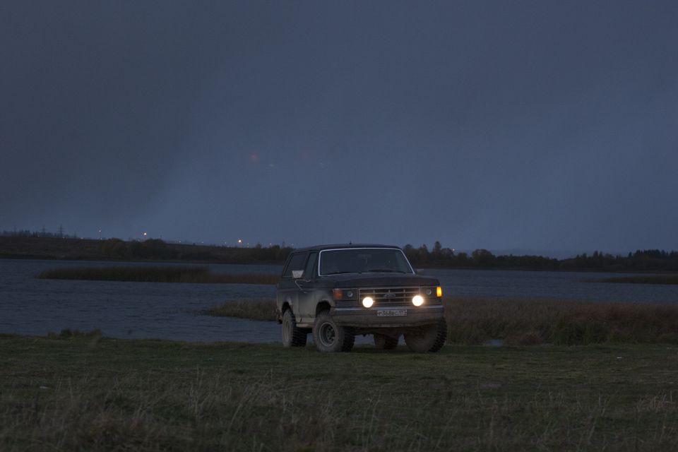 1987 Ford Bronco driving on a dirt road at dusk, headlights on