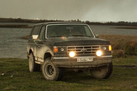 1987 Ford Bronco - A 1987 Ford Bronco is shown from the front, parked on grassy terrain next to a body of water.