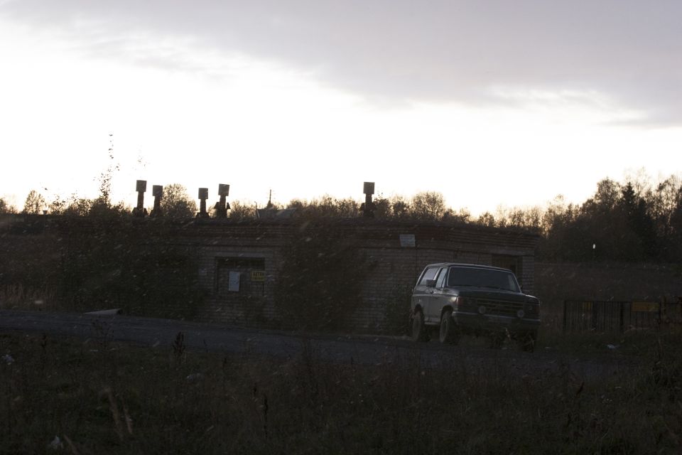 1987 Ford Bronco parked on a dirt road in front of a brick building with multiple chimneys