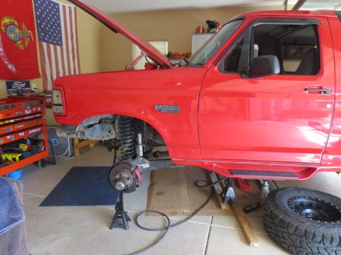 1996 Ford Bronco - Front end of a red Ford Bronco is raised on jack stands in a garage, undergoing maintenance.
