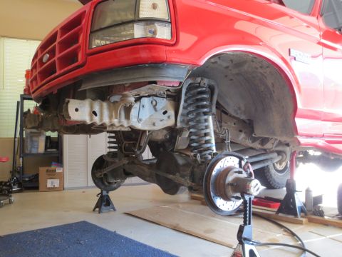 1996 Ford Bronco - Front suspension of a red Ford truck is visible with the bumper removed and the vehicle supported by jack stands.