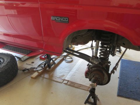1996 Ford Bronco - The front suspension of a red Ford Bronco is undergoing maintenance, with the wheel off and the vehicle elevated on jack stands.