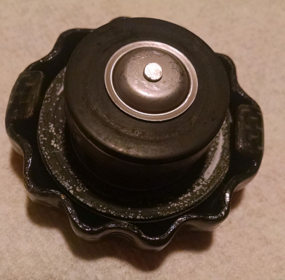 Radiator cap with a metal center and black rubber seal. Some white residue is visible on the black metal base