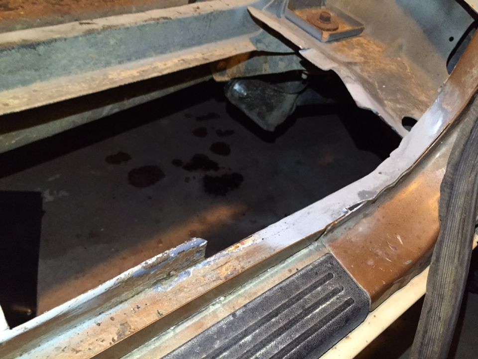 1991 Ford Bronco floor pan with significant rust and holes, showing a dark, wet surface below