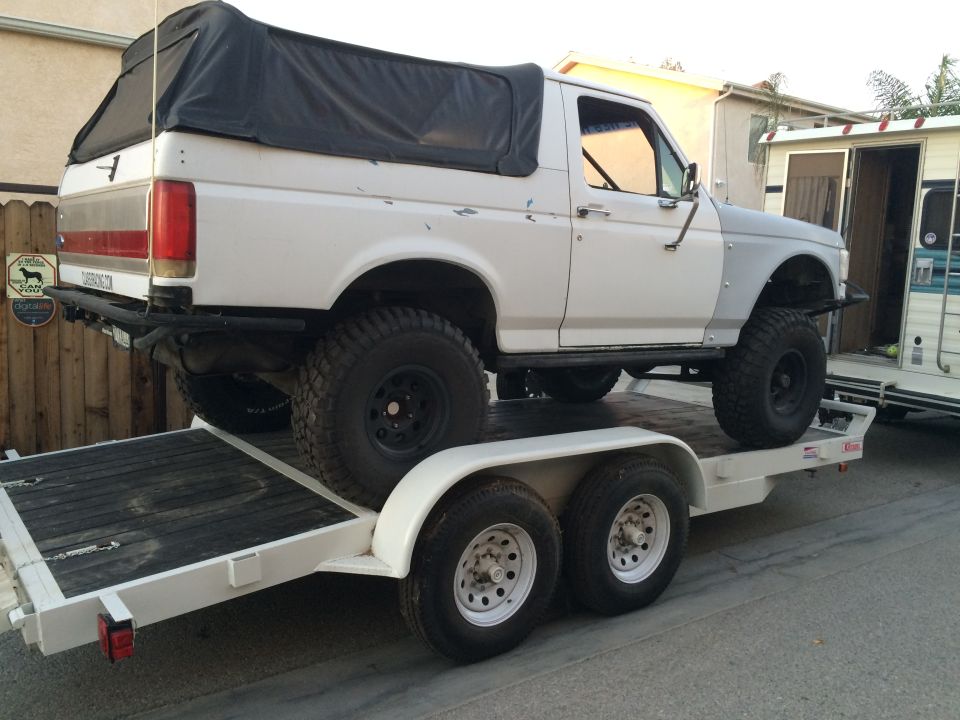 White Ford Bronco with black soft top on a dual-axle trailer, gray fender flares, off-road tires