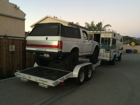 1997 Dodge 3500 - A lifted white Bronco is transported on a trailer, towed by a pickup truck with a camper shell.