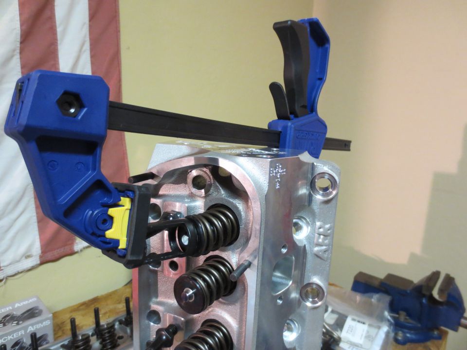 Engine cylinder head held by a clamp, showing valve springs and ports. Text visible on the part