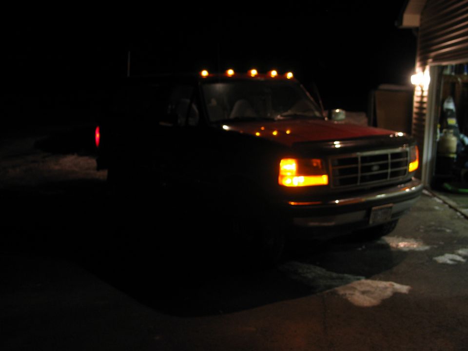 Dark Ford truck at night with headlights and roof marker lights on, beside a lit garage.
