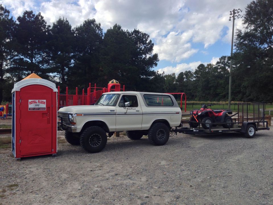 White Ford Bronco towing a trailer with a red ATV. Portable toilet is to the left